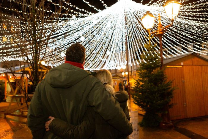 Eine Aufnahme eines Pärchens von hinten, dick bekleidet, auf dem Weihnachtsmarkt in Ettelbruck. Sie schauen sich die umliegenden Stände und die schöne Lichtinstallation an.